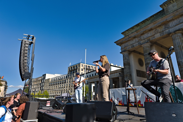 Eine Gruppe von Menschen, die auf einer Bühne Musik spielen, mit dem Brandenburger Tor im Hintergrund, begleitet von Equipment und einem klaren Himmel.