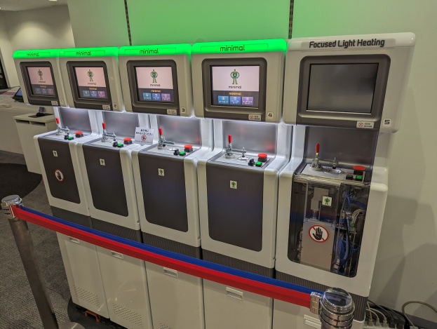 Eine Reihe von Mini-Heizungssystemgeräten mit Knöpfen und Drehreglern, die nebeneinander in einem Raum mit einer Wand im Hintergrund, Deckenlampen darüber und einem Boden darunter angeordnet sind.