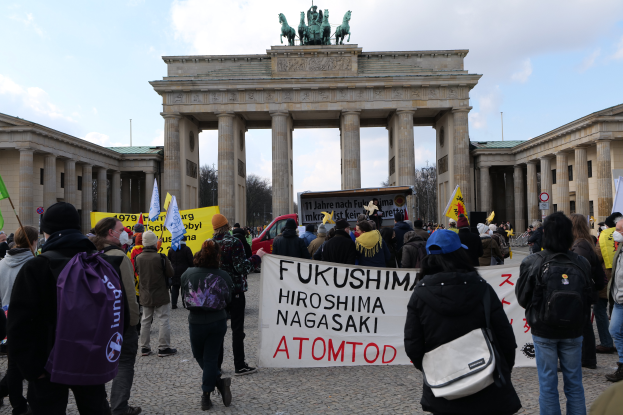 Eine Gruppe von Menschen steht vor dem Brandenburger Tor in Berlin, Deutschland, mit Schildern und Fahnen, auf denen "Fukushima Hiroshima Nagasaki Atomod" steht, um gegen die Atombombe zu protestieren, wobei das Tor, seine Bögen, Säulen, Statuen, Bäume und Fahrzeuge im Hintergrund zu sehen sind.