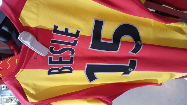 Ein roter und gelber Fußballtrikot mit Text und Zahlen hängt an einer Stange in einem Geschäft, mit anderen Gegenständen und dem Boden im Hintergrund.