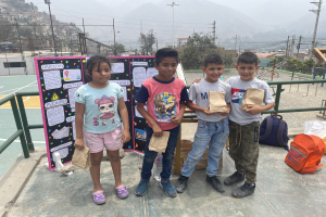 Gruppe von Kindern auf einem Basketballfeld mit Papierbeuteln in den Händen, Beutel und Gegenstände rechts und eine Tafel mit Text links, vor einem Hintergrund aus Zaun, Pfählen, Straßenlaternen, Bäumen, Gebäuden, Hügeln und Himmel.