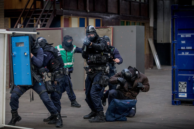 Polizeibeamte in Einsatzausrüstung stehen vor einem Gebäude, einige halten Waffen, mit einer Person am Boden im Vordergrund und metallischen Gegenständen, Treppen und einer Wand im Hintergrund.