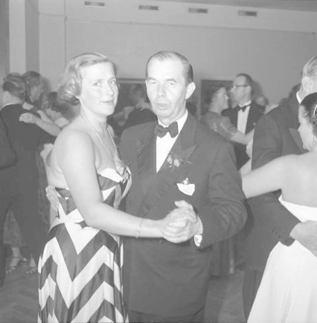 Ein Mann in einem schwarzen Blazer, weißem Hemd und schwarzen Hosen tanzt mit einer Frau in einem unbekannten Outfit auf einer formellen Veranstaltung, indem sie sich an den Händen halten und zur Musik wiegen.