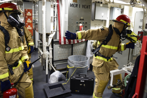 Drei Feuerwehrleute in Schutzausrüstung stehen zusammen in einem Raum mit einem Feuerlöscher links, einer Maschine rechts und diversen Gegenständen im Hintergrund.