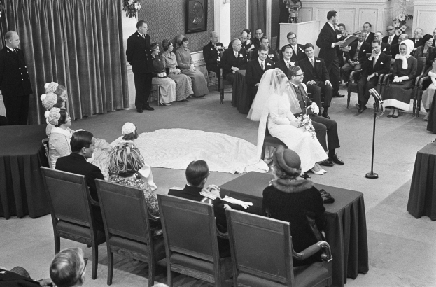 Schwarzes und weißes Foto einer Hochzeitszeremonie mit sitzenden und stehenden Gästen, einem Mann mit Mikrofon, einer Frau mit einem Bukett, einem Podium mit Mikrofon, Blumenschmuck, einem Vorhang, einem gerahmten Foto an der Wand und einer Tür im Hintergrund.