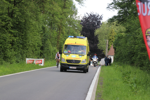 Ambulanz fährt auf einer Straße mit Radfahrern daneben, Gras und Bäume auf beiden Seiten, Häuser und Masten im Hintergrund unter einem klaren blauen Himmel.