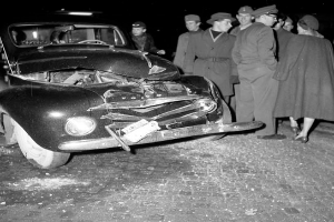 Ein Schwarz-Weiß-Bild eines verunglückten Autos am Straßenrand mit eingedellter Motorhaube und zersplitterten Fenstern, umgeben von einer Gruppe besorgter Menschen.