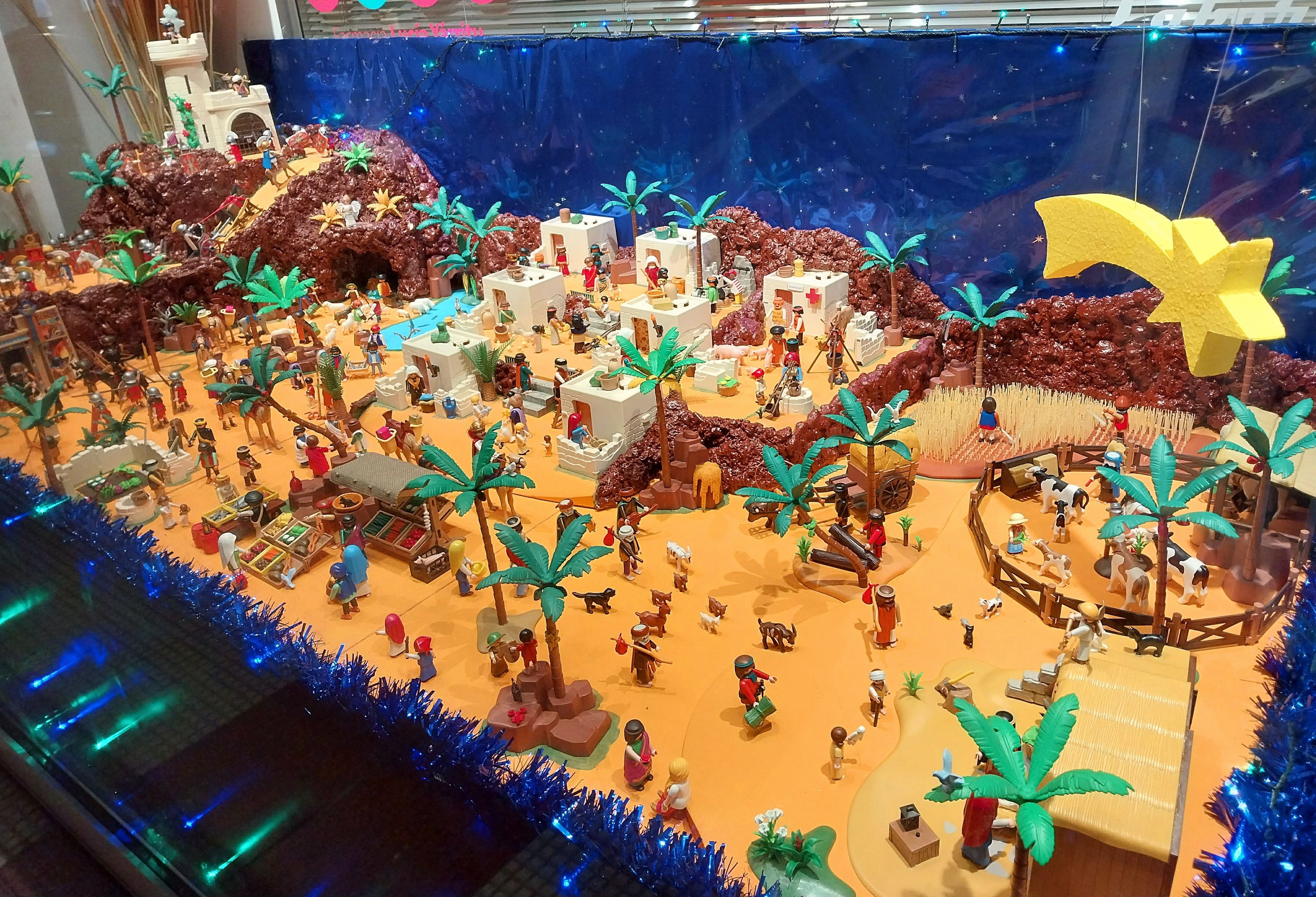 Vitrine voller verschiedener Lego-Steine auf einem Tisch, umgeben von Spielzeugen, Bäumen und dekorativen Gegenständen, mit einer festlich beleuchteten Wand im Hintergrund.