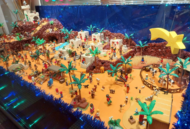 Vitrine voller verschiedener Lego-Steine auf einem Tisch, umgeben von Spielzeugen, Bäumen und dekorativen Gegenständen, mit einer festlich beleuchteten Wand im Hintergrund.
