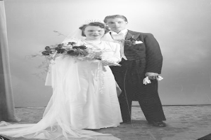 Ein Mann und eine Frau in festlicher Kleidung stehen auf einer Bühne mit einem Vorhang und einer Wand dahinter, die Frau hält einen Blumenstrauß und der Mann hält ein Objekt, auf einem Schwarz-Weiß-Bild.