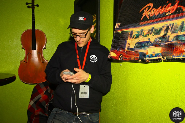Eine Person mit einer Mütze, die ein Handy hält, trägt eine ID-Karte und hat links eine Gitarre und rechts ein Plakat
