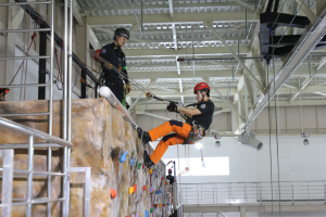 Zwei Personen in schwarzer und oranger Kleidung und Helmen klettern an einer Felswand in einer Halle, gesichert durch Seile, mit einem Metallgeländer links und einer weißen Wand mit Leuchten im Hintergrund.
