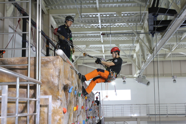 Zwei Personen in schwarzer und oranger Kleidung und Helmen klettern an einer Felswand in einer Halle, gesichert durch Seile, mit einem Metallgeländer links und einer weißen Wand mit Leuchten im Hintergrund.