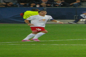 Ein Mann in weißem Hemd und roter Shorts, der auf einem Feld Fußball spielt, umgeben von einer Gruppe von Menschen im Hintergrund.