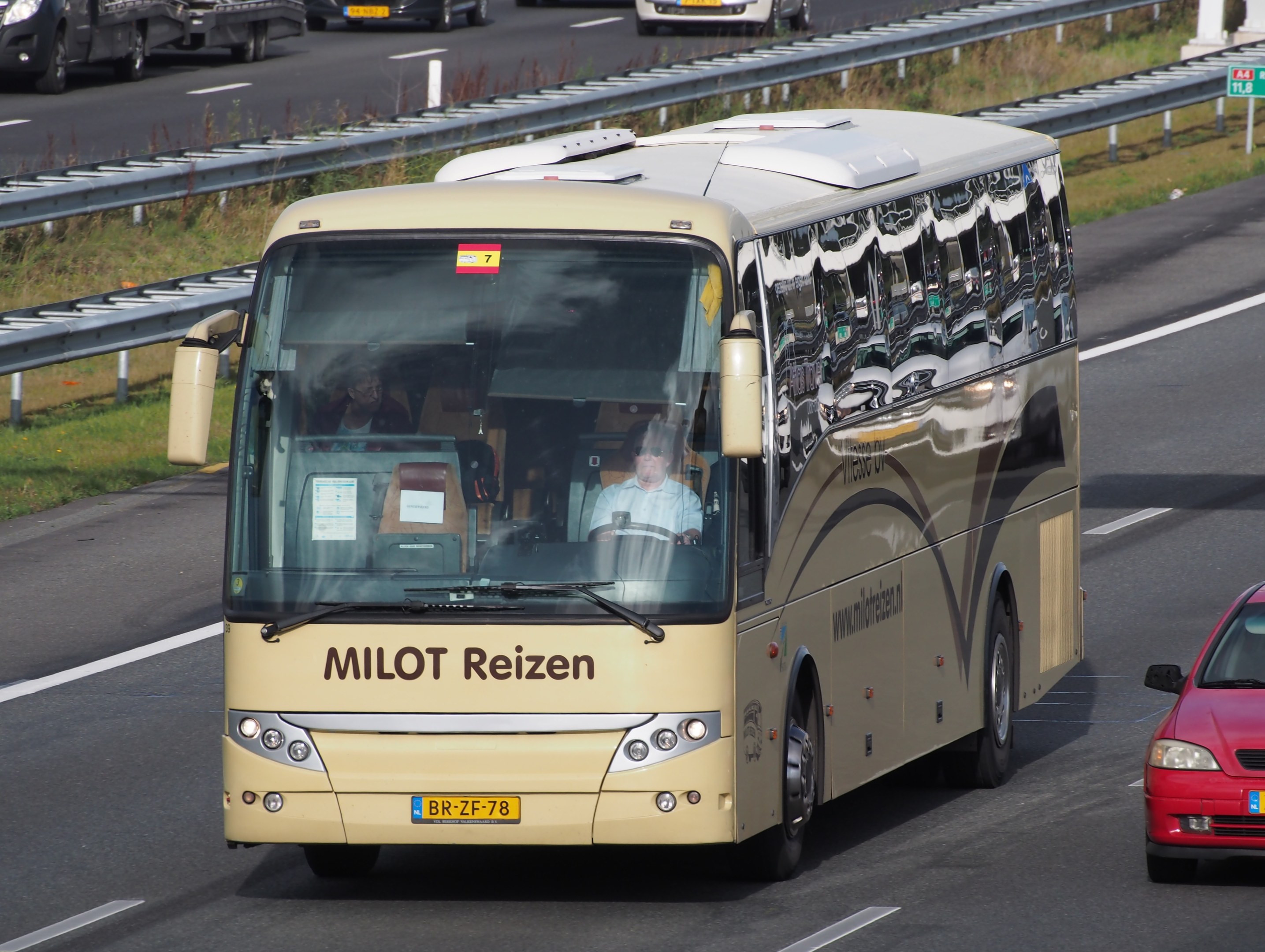Ein Minibus mit Passagieren auf einer Autobahn neben einem roten Auto, mit Leitplanken und Gras an den Seiten und Texttafeln und Säulen im Hintergrund.