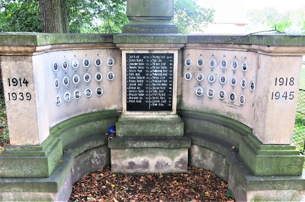 Ein Holocaust-Gedenkmonument in einem Berliner jüdischen Friedhof, mit einer Text- und Nummerntafel an seiner Wand, umgeben von Bäumen, einem Zaun und verstreuten trockenen Blättern.