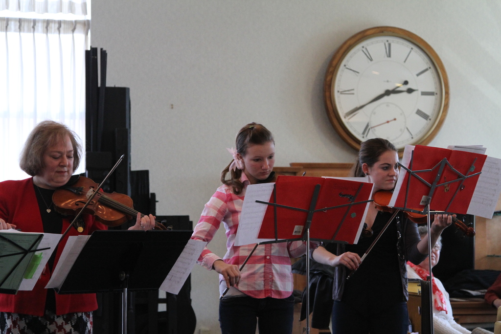 Drei Frauen auf der Bühne, die Geigen spielen, mit Notenpulten und Noten davor, während ein Zuschauer im Vordergrund sitzt; eine Uhr hängt an der weißen Wand im Hintergrund, und ein weißer Vorhang ist links sichtbar.