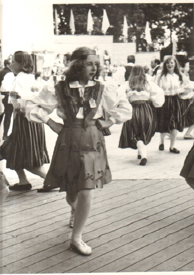 Eine Gruppe von Frauen in traditioneller bayrischer Tracht tanzt auf einem Holzboden, mit einer Wand, Bäumen und anderen Gegenständen im Hintergrund; Text ist am unteren Bildrand vorhanden.