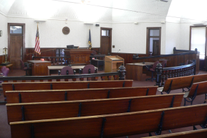 Ein Gerichtssaal mit hölzernen Kirchenbänken, Tischen, Stühlen, einem Podium, Flaggen, Türen, einer Uhr an der Wand und Lampen und Ventilatoren an der Decke.