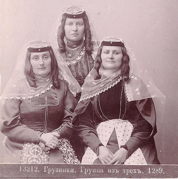 Schwarz-weißes Foto von drei Frauen in traditionellen russischen Hochzeitskleidern mit farbenfrohen Stickereien und Perlen gegen eine einfarbige weiße Wand.