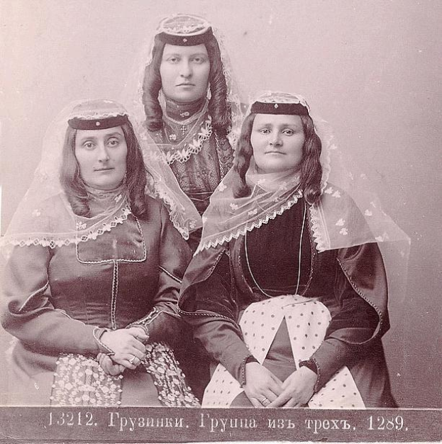 Schwarz-weißes Foto von drei Frauen in traditionellen russischen Hochzeitskleidern mit farbenfrohen Stickereien und Perlen gegen eine einfarbige weiße Wand.
