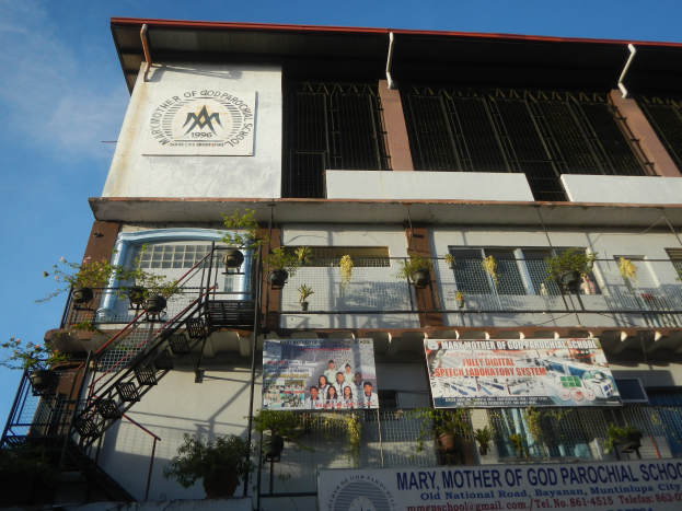 Mariä Himmelfahrt Pfarrschule Gebäude mit Fenstern, Geländern, Treppen, Topfpflanzen, Banner, Wolkenhimmel im Hintergrund.