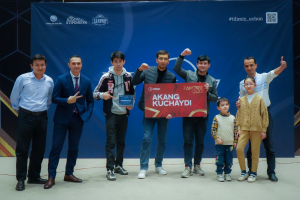 Eine Gruppe von Menschen auf einer Bühne, zwei halten eine Tafel mit der Aufschrift "Akang Kuchaydi - Zakovat Quiz", mit einem Banner, Lichtern und anderen Gegenständen im Hintergrund.