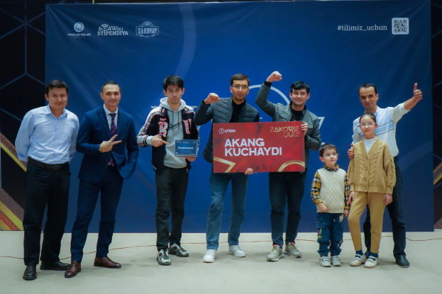 Eine Gruppe von Menschen auf einer Bühne, zwei halten eine Tafel mit der Aufschrift "Akang Kuchaydi - Zakovat Quiz", mit einem Banner, Lichtern und anderen Gegenständen im Hintergrund.
