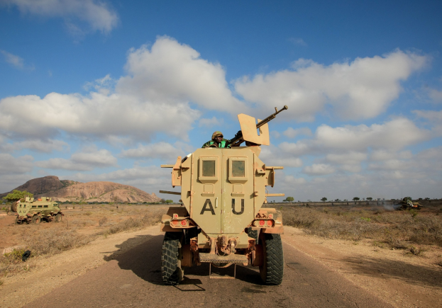Militärisches gepanzertes Mannschaftsfahrzeug auf einer Schotterstraße durch einen bewaldeten Bereich mit einem bewölkten Himmel im Hintergrund, zwei Insassen sind im Inneren sichtbar.