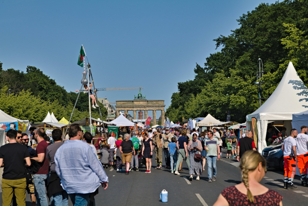 Menschenmenge geht eine Straße entlang mit Zelten, Fahrzeugen und Bäumen bei einem Event mit Fahnenmasten, einem Bogen und einem klaren blauen Himmel.