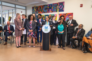 Gruppe von Menschen bei einer Stadtratssitzung mit einem Podium, Mikrofon und einer Tafel, die 'Sancho Stadtrat tritt dem Stadtrat bei' trägt, umgeben von sitzenden und stehenden Zuschauern, Flaggen und einem Feuerlöscher.