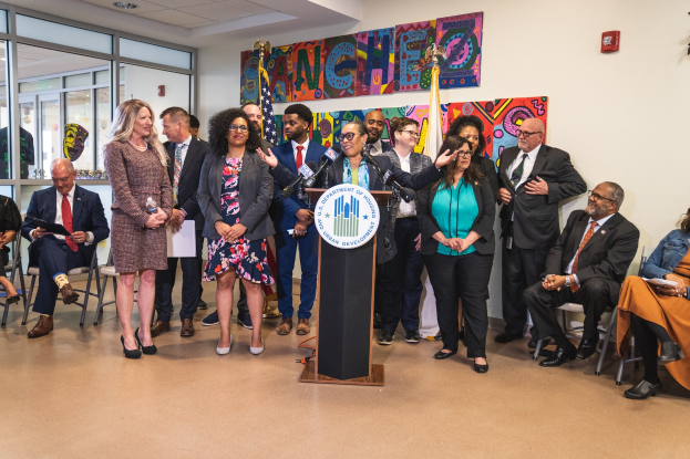 Gruppe von Menschen bei einer Stadtratssitzung mit einem Podium, Mikrofon und einer Tafel, die 'Sancho Stadtrat tritt dem Stadtrat bei' trägt, umgeben von sitzenden und stehenden Zuschauern, Flaggen und einem Feuerlöscher.