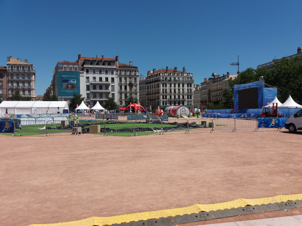 Großer städtischer Platz voller Zelte, umgeben von Gebäuden, Bäumen, Fahrzeugen auf der Straße und einem Geländer unter einem klaren blauen Himmel.
