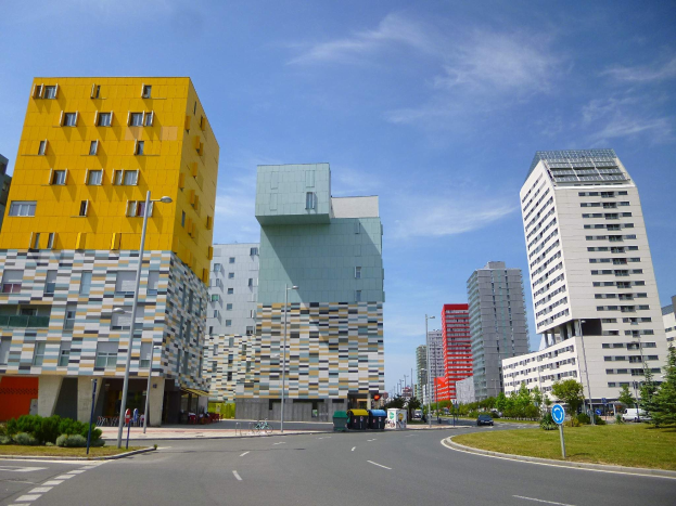 Eine Stadtstraße mit hohen Gebäuden im Hintergrund, einschließlich Strukturen der Universität Rotterdam, Straßenlaternen, Straßenschildern, Motorfahrzeugen, Informationsanzeigen, Büschen, Bäumen und einem bewölkten Himmel.