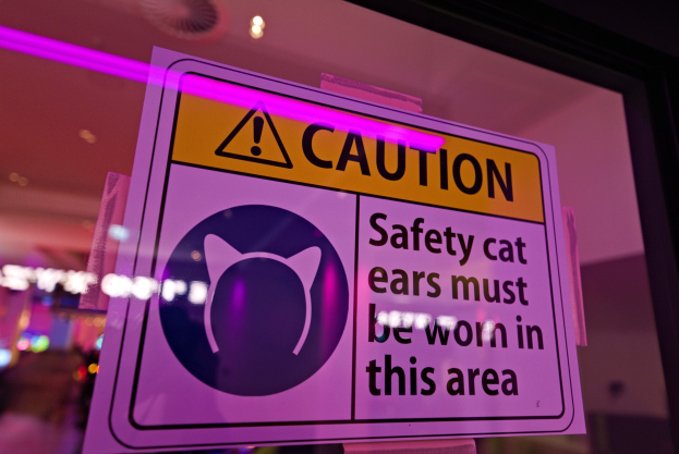 Vorsichtsschild in einem Schaufenster mit der Aufschrift "Vorsicht: Sicherheitskatzenohren müssen in diesem Bereich getragen werden" auf einem bunten Hintergrund.