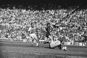 Schwarzes Foto von Männern beim Fußballspielen auf einem Feld mit Zuschauern im Hintergrund und einem Banner "Bundesliga 1978-79 Ajax 1-1 Hannover 96".