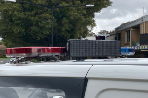 Ein Polizeiwagen mit eingeschaltetem Blaulicht steht am Straßenrand, umgeben von Gebäuden, Bäumen, Pfählen und Schildern mit Text, unter einem bewölkten Himmel, wahrscheinlich nach einem tödlichen Schusswaffengebrauch an einer Schule in Brisbane.