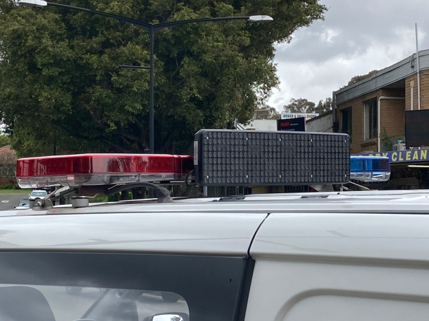 Ein Polizeiwagen mit eingeschaltetem Blaulicht steht am Straßenrand, umgeben von Gebäuden, Bäumen, Pfählen und Schildern mit Text, unter einem bewölkten Himmel, wahrscheinlich nach einem tödlichen Schusswaffengebrauch an einer Schule in Brisbane.