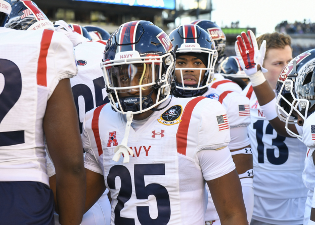 Gruppe von Männern in Football-Uniformen auf einem Feld mit einem unscharfen Publikum, Scoreboard und Himmel im Hintergrund.