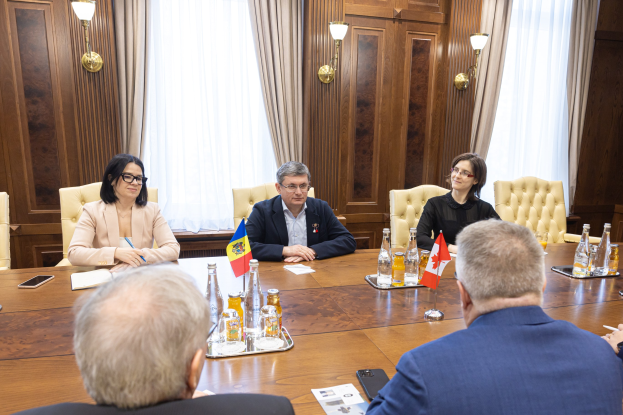 Eine Gruppe von Menschen, die um einen hölzernen Tisch in einer förmlichen Umgebung sitzen und sich mit EU-bezogenen Materialien beschäftigen, umgeben von Flaggen, Flaschen, Gläsern, Papieren und Mobiltelefonen.