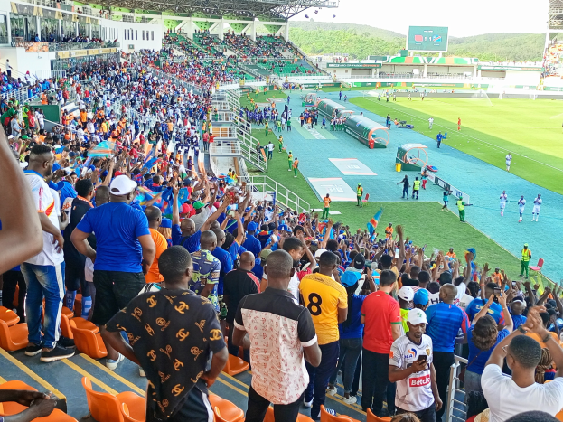 Eine große Menschenmenge versammelt sich vor einem eingezäunten Fußballfeld mit Hügeln und einem blauen Himmel im Hintergrund und erwartet ein Spiel während der Afrikameisterschaft 2019.