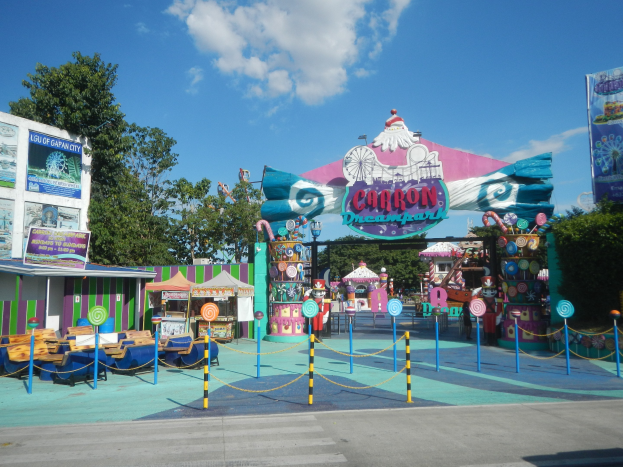 Kirmesfahrgeschäft am Parkeingang mit Straße, eingezäuntem Bereich, Buden, Schildern, Bäumen und bewölktem Himmel im Hintergrund.