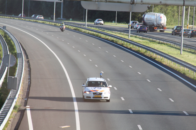 Ein Auto auf einer Autobahn mit einem stationären Polizeiwagen am Straßenrand, andere Fahrzeuge, Leitplanken, Strommasten, Verkehrsschilder und Bäume im Hintergrund.