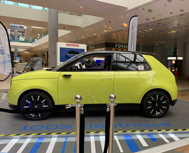 Ein gelbes wasserstoffbetriebenes Elektroauto, das drinnen auf einem Parkplatz steht und von Absperrpollern mit Seilen, Bannern und Schildern umgeben ist, mit einer Person im Auto und einer Gruppe von Menschen in der Nähe.