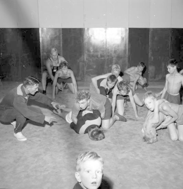 Schwarz-weißes Bild von jungen Jungs, die in einer Turnhalle miteinander ringen, mit einer Wand im Hintergrund.
