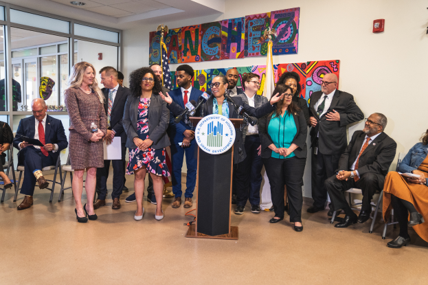 Eine Gruppe von Menschen, die vor einem Podium mit Mikrofon und einer Tafel mit der Aufschrift "Sancho Stadtrat tritt dem Stadtrat bei" stehen und sitzen, mit Flaggen, Feuerlöscher und Deckenleuchten im Hintergrund.