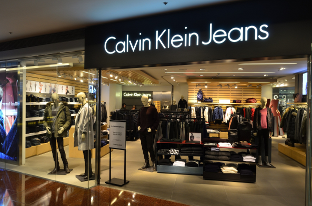 Innenansicht eines Calvin Klein Jeans-Geschäfts in einem Einkaufszentrum, mit Schaufensterpuppen in modischen Outfits, aufgehängten Kleidungsstücken, einem Werbeplakat, einem digitalen Bildschirm und Deckenbeleuchtung.