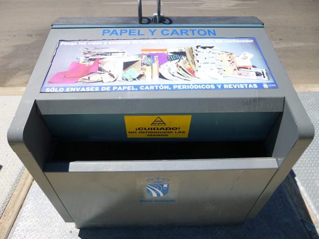 Ein Metallmüllcontainer am Straßenrand mit einem "Papel y Carton"-Plakat, mit im Hintergrund fahrenden Fahrzeugen.