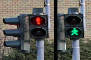 Zwei Ampeln an einem Mast vor einer Backsteinwand mit Bäumen im Hintergrund, die eine rote und grüne Lampe anzeigen, die den Überqueren der Straße durch eine Person anzeigen.