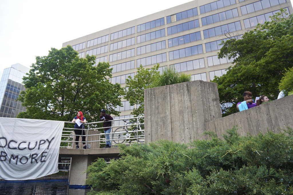 Menschen auf einer Brücke mit einem Banner und einem Fahrrad, mit Wasser darunter und Bäumen, Gebäuden und Himmel im Hintergrund.